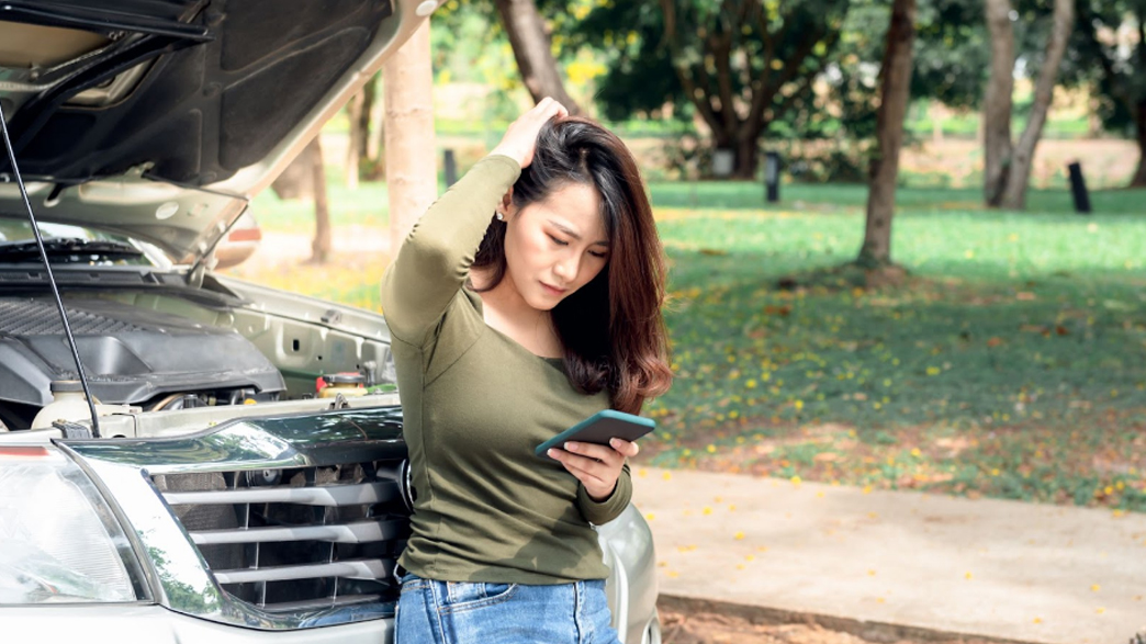 A stranded woman stands on the side of the road after her vehicle breaks down, calling for help.