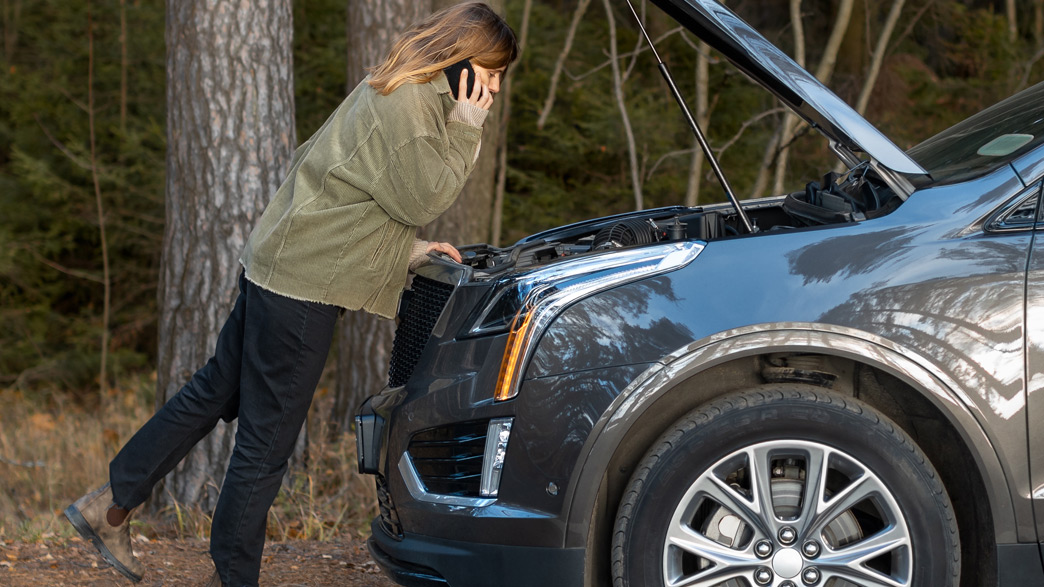 Woman on forest road, on the phone with car hood open after potential lifter failure leaves her stranded with engine trouble.