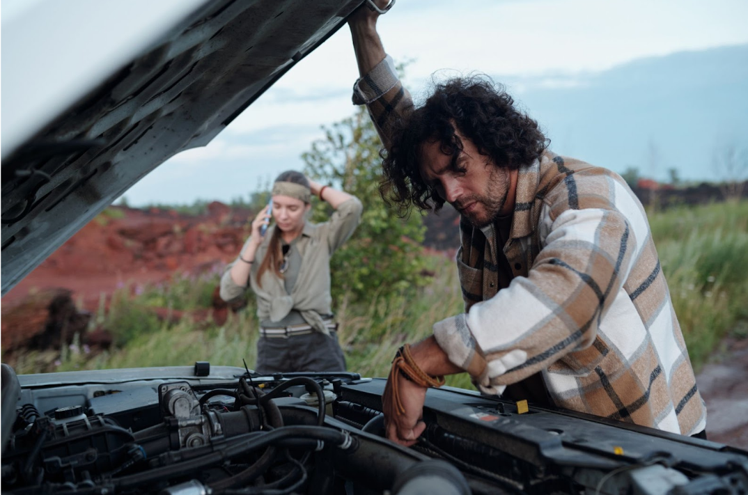 Man inspecting engine while woman calls for help after engine failure on rural roadside.