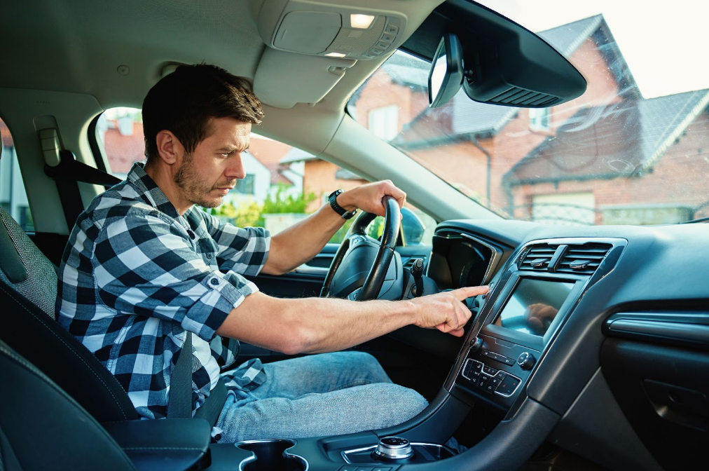 Man in plaid shirt parked in driveway, uses car touchscreen, possibly checking an issue with vehicle engine noise at startup.